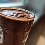 Close-up, sunlit view of a thick, blended frozen hot chocolate in a tall glass, showing condensation.