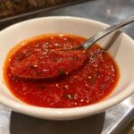 A spoonful of vibrant red, chunky pizza sauce with visible herbs and seeds being lifted from a small white bowl.