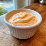 Close-up of a creamy, orange-hued burger sauce speckled with red spices, served in a small white ramekin.