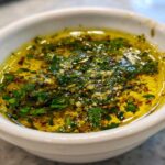 Close-up of flavorful olive oil bread dip with minced garlic and fresh green herbs in a white bowl.