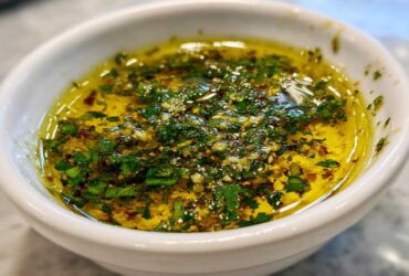 Close-up of flavorful olive oil bread dip with minced garlic and fresh green herbs in a white bowl.