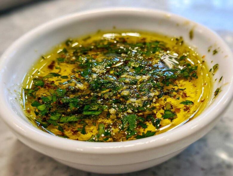 Close-up of flavorful olive oil bread dip with minced garlic and fresh green herbs in a white bowl.