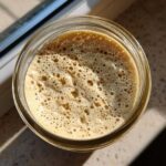 Overhead view of a bubbly, active sourdough starter in a glass jar, showing signs of fermentation.