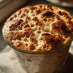Close-up of a very active sourdough starter that has risen and overflowed slightly, showing a bubbly, browned top in a clear glass jar.