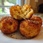 Four golden brown, deep-fried rice balls, one cut open showing melted cheese and macaroni filling.