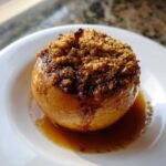 A close-up of a single, glistening baked apples dessert topped with a brown sugar crumble and sitting in a pool of syrup on a white plate.