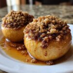 Two glistening baked apples topped with a brown sugar and oat crumble, sitting in sweet syrup on a white plate.