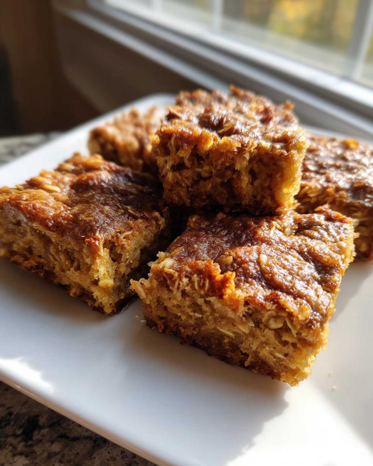 Close-up of golden brown squares of baked oatmeal stacked on a white plate near a window.