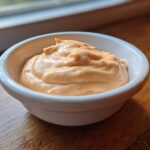 A close-up of creamy, orange-tinted burger sauce served in a small white ramekin on a wooden surface.