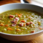 Close-up of a white bowl filled with thick, green pea soup topped with chunks of ham and diced carrots.