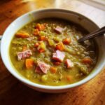 Close-up of a thick, green pea soup topped with diced ham, carrots, and black pepper, served in a white bowl.