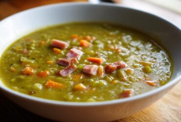 Close-up of a white bowl filled with thick, green pea soup topped with chunks of ham and diced carrots.