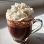 A clear glass mug filled with rich dark chocolate drink topped high with fluffy whipped hot chocolate.
