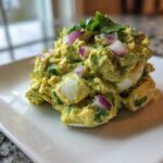 Close-up of a mound of creamy avocado egg salad mixed with chopped red onion and cilantro, served on a white plate.