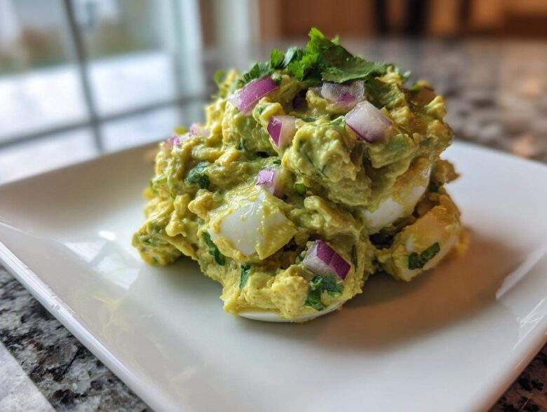 Close-up of a mound of creamy avocado egg salad mixed with chopped red onion and cilantro, served on a white plate.