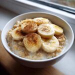 A white bowl filled with creamy banana oatmeal, topped with sliced bananas and a dusting of cinnamon.