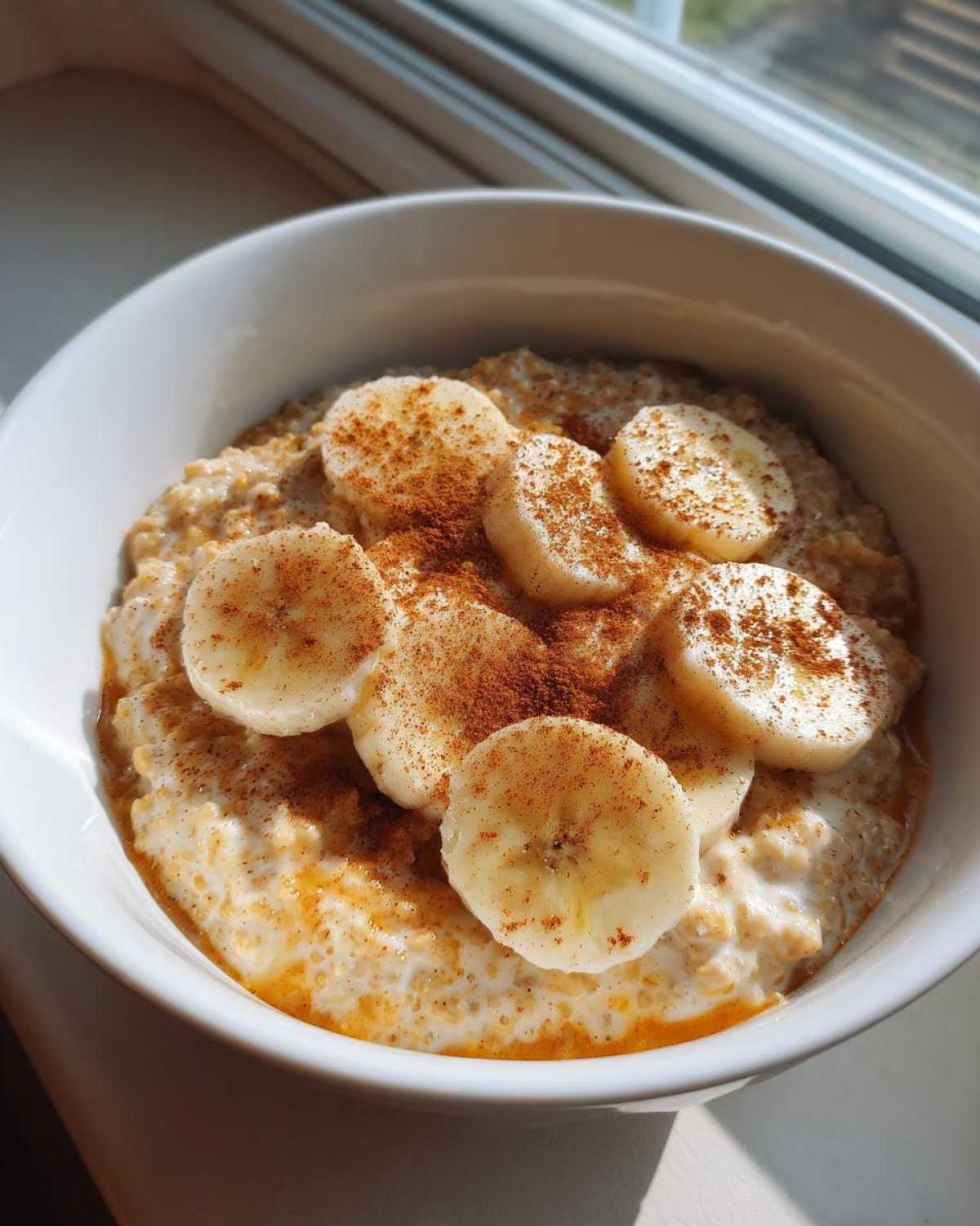 A white bowl filled with creamy banana oatmeal, topped with sliced bananas and a generous dusting of cinnamon.