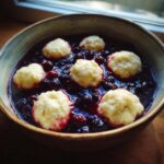 Six fluffy, light-colored Blackberry Dumplings served in a rich, dark purple blackberry sauce in a rustic bowl.