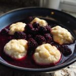 A close-up of fluffy dumplings served in a rich, dark purple sauce with whole fresh blackberries, showcasing the Blackberry Dumplings.