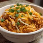 A close-up of a white bowl filled with creamy buffalo chicken chili, topped with shredded cheddar and Monterey Jack cheese and green onions.