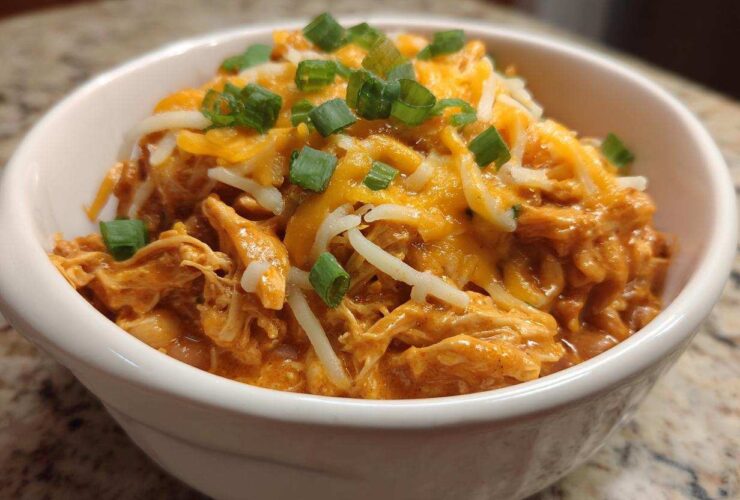 A close-up of a white bowl filled with creamy buffalo chicken chili, topped with shredded cheddar and Monterey Jack cheese and green onions.