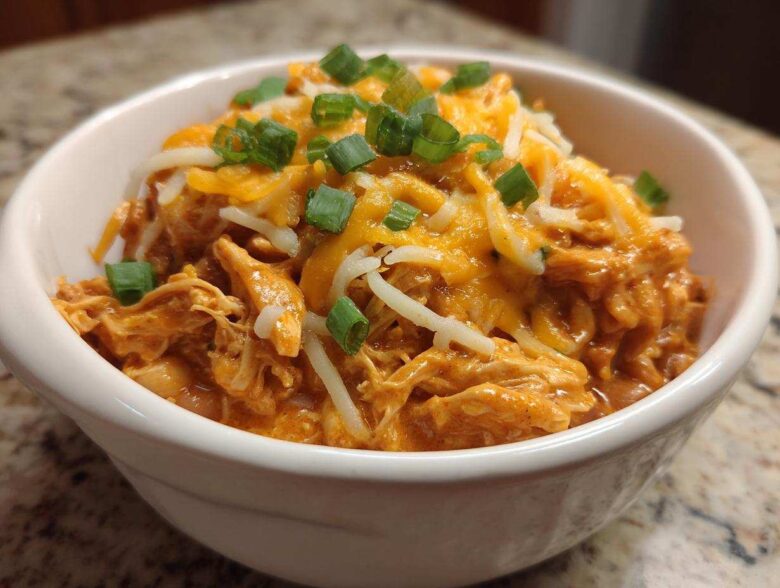 A close-up of a white bowl filled with creamy buffalo chicken chili, topped with shredded cheddar and Monterey Jack cheese and green onions.