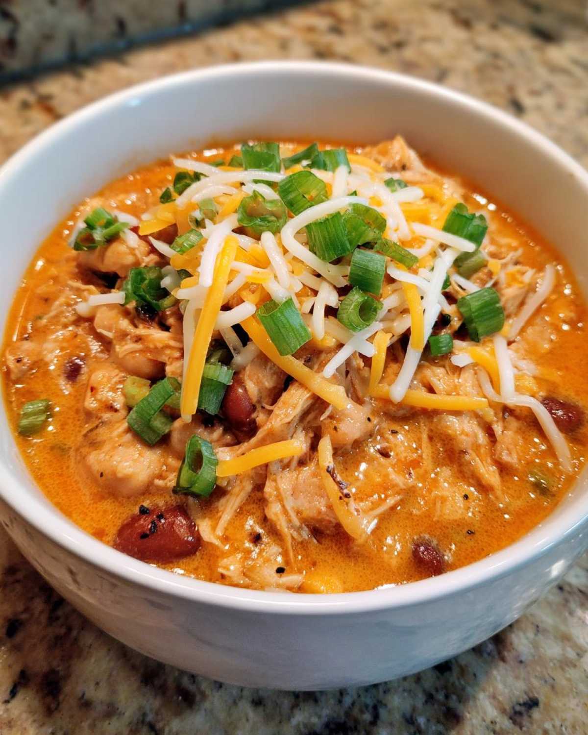 Close-up of a white bowl filled with creamy buffalo chicken chili, topped with shredded cheese and green onions.