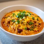 Close-up of a bowl of creamy buffalo chicken chili topped with shredded cheddar cheese and sliced green onions.