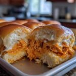A close-up of a buffalo chicken sliders sandwich cut in half, showing shredded chicken coated in orange sauce and melted white cheese.