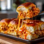 A serving utensil pulls apart a cheesy, saucy buffalo chicken slider, showing long cheese pulls.