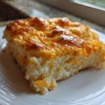 A square serving of cheesy baked mashed potatoes with a golden-brown crust, served on a white plate.
