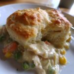 A close-up of a serving of creamy chicken pot pie casserole topped with a golden, fluffy biscuit.