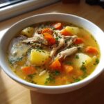 A close-up of a white bowl filled with rich, golden chicken vegetable soup, featuring shredded chicken, chunks of potato, and bright carrots.