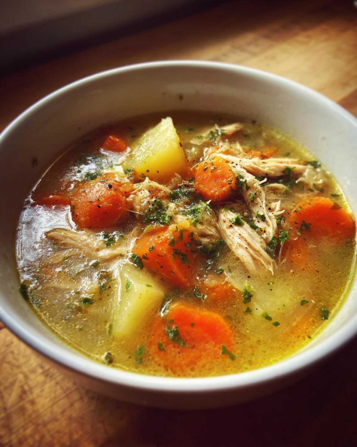 Close-up of a white bowl filled with steaming chicken vegetable soup, featuring shredded chicken, carrots, and potatoes.