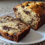 A close-up of a freshly baked chocolate chip banana bread recipe loaf, partially sliced on a white plate.