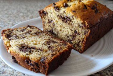 A close-up of a freshly baked chocolate chip banana bread recipe loaf, partially sliced on a white plate.