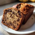 Close-up of a moist slice of chocolate chip banana bread next to the loaf on a white plate.