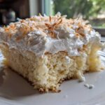 A close-up of a moist slice of coconut poke cake topped with whipped cream and toasted coconut flakes.