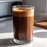 A tall glass filled with a cold, dark brown Coffee Smoothie topped with foam, showing condensation.