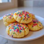 A stack of soft, pale yellow cake batter cookies generously topped with bright, multi-colored sprinkles.