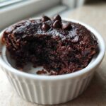 A close-up of a rich, dark chocolate Cottage Cheese Microwave Brownie in a white fluted ramekin, topped with chocolate chips.
