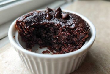 A close-up of a rich, dark chocolate Cottage Cheese Microwave Brownie in a white fluted ramekin, topped with chocolate chips.