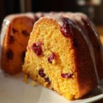 Close-up of a slice of moist cranberry orange cake showing dried cranberries embedded in the yellow crumb and topped with a white glaze.