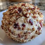 A close-up of a perfectly formed cranberry pecan cheese ball coated in chopped pecans and dried cranberries.