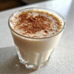 Close-up of a glass filled with creamy coquito recipe, topped with a generous dusting of ground cinnamon.