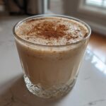 A glass of creamy, frothy coquito recipe drink dusted heavily with ground cinnamon.