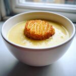 A bowl of creamy yellow garlic soup topped with a large, golden-brown, ridged crouton.