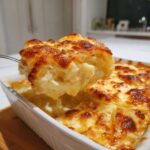 A spoonful of creamy scalloped potatoes is lifted from a white baking dish showing a golden, bubbly, and browned cheese crust.