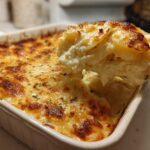 A scoop of creamy scalloped potatoes being lifted from a baking dish showing a golden-brown cheesy crust.
