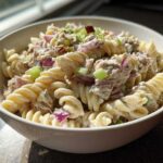 A close-up of creamy Tuna pasta salad made with rotini pasta, tuna, red onion, and celery in a white bowl.
